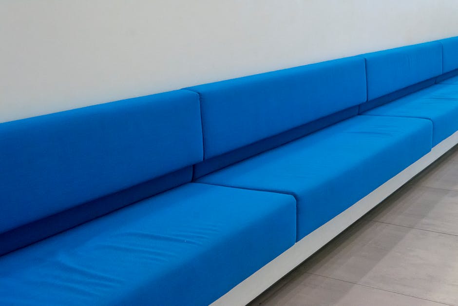 A long, bright blue upholstered bench with a smooth surface, positioned against a plain white wall in a modern, well-lit interior space. The bench consists of multiple connected seat cushions with min